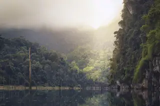 Ilustrasi: Danau berair tenang di antara bukit berhutan lebat yang diselimuti kabut dan awan.