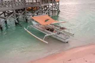 Ilustrasi: Sebuah perahu kayu bercadik dengan atap terpal oranye bersandar di tepi pantai dekat dermaga kayu.