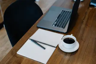 Ilustrasi: Buku catatan spiral, pena, secangkir kopi hitam, dan laptop terbuka di atas meja kayu.