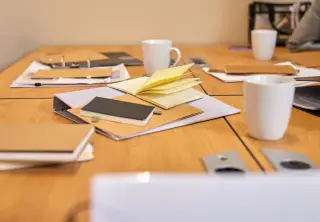 Ilustrasi: Buku catatan terbuka, map dokumen, dan cangkir kopi di atas meja rapat kayu.