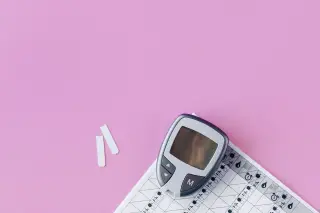 Ilustrasi: Alat glukometer dan strip tes berada di atas catatan medis dengan latar belakang merah muda.
