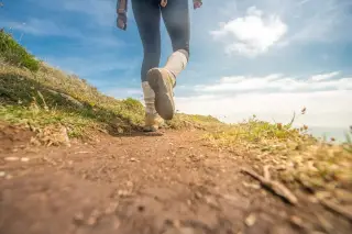 Ilustrasi: Seseorang mengenakan sepatu trekking berjalan di jalur setapak alam berumput dengan latar langit biru.