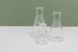 Ilustrasi: Tiga labu erlenmeyer kaca kosong di atas meja putih dengan latar belakang hijau.