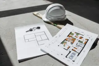 Ilustrasi: Denah arsitektur, helm proyek, alat pengukur kedataran, dan kunci tergeletak di lantai.