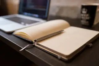 Ilustrasi: Buku catatan terbuka dengan pena di atas meja dekat laptop dan cangkir.