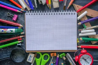 Ilustrasi: Buku catatan bergaris kotak dikelilingi berbagai perlengkapan sekolah dan alat tulis.