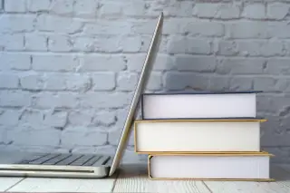 Ilustrasi: Sebuah laptop terbuka di samping tumpukan tiga buku di atas meja kayu dengan latar dinding bata.