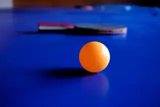 Ilustrasi: Sebuah bola tenis meja oranye dan bet di atas permukaan meja berwarna biru.