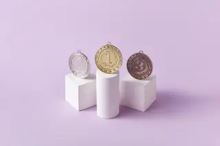 Ilustrasi: Tiga medali bernomor 1, 2, dan 3 di atas podium putih dengan latar belakang ungu muda.