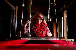 Ilustrasi: Seorang wanita berkerudung merah sedang menenun kain tradisional dengan alat tenun kayu.
