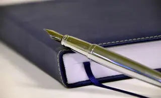 Ilustrasi: Sebuah pena bertinta perak diletakkan di atas buku catatan bersampul biru tua.