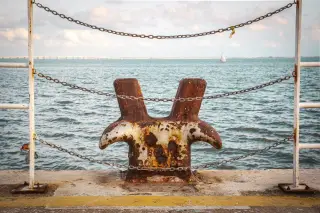 Ilustrasi: Bollard dermaga berkarat dibatasi rantai dengan latar belakang laut dan perahu layar.