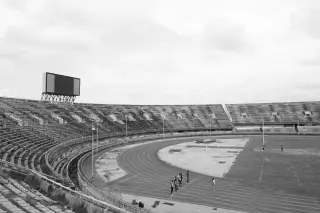 Ilustrasi: Foto hitam putih sebuah stadion olahraga besar dengan lintasan lari dan beberapa orang di tepinya.