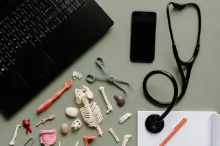 Ilustrasi: Stetoskop, model anatomi tulang mini, laptop, dan ponsel di atas meja.
