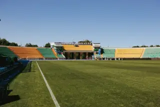 Ilustrasi: Pemandangan lapangan sepak bola dan tribun stadion dengan kursi kuning dan hijau di bawah langit cerah.