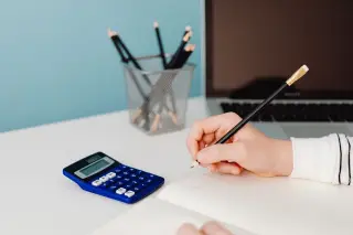 Ilustrasi: Tangan seseorang menulis dengan pensil di buku dekat kalkulator dan laptop.