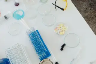 Ilustrasi: Peralatan laboratorium, tabung reaksi, cawan petri, dan obat-obatan di atas meja putih.