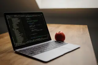 Ilustrasi: Sebuah laptop terbuka menampilkan kode pemrograman di samping buah apel merah di atas meja kayu.