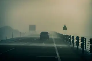Ilustrasi: Sebuah mobil melintas di jalan raya beraspal yang diselimuti kabut tebal dengan jarak pandang rendah.