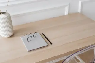 Ilustrasi: Buku catatan kecil, kacamata, pensil, dan vas di atas meja kayu minimalis.