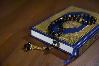 Ilustrasi: Sebuah kitab suci bersampul biru dan untaian tasbih hitam di atas meja kayu.
