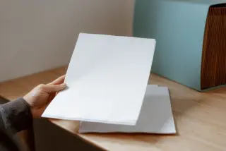 Ilustrasi: Tangan memegang tumpukan lembaran kertas putih kosong di atas meja kayu dekat map berkas.