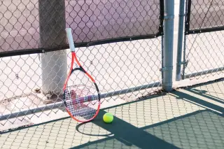 Ilustrasi: Sebuah raket dan bola tenis diletakkan di tepi lapangan dekat pagar kawat di bawah terik matahari.