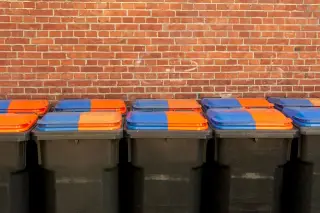 Ilustrasi: Deretan tempat sampah beroda dengan penutup oranye dan biru di depan dinding bata merah.
