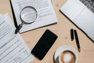 Ilustrasi: Tampak atas meja kerja berisi dokumen berkas, kaca pembesar, laptop, ponsel, dan secangkir kopi.