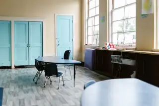 Ilustrasi: Ruang kelas kosong dengan meja bundar biru, kursi, dan pintu kayu berwarna biru muda.