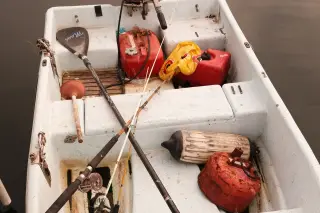 Ilustrasi: Tampak atas perahu motor kecil berisi jeriken bahan bakar merah, dayung, dan alat pancing.