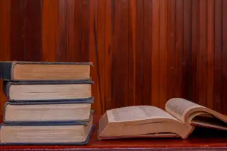 Ilustrasi: Tumpukan buku tebal dan satu buku terbuka di atas meja kayu dengan latar dinding kayu.