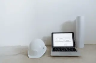 Ilustrasi: Helm proyek putih, laptop dengan denah bangunan, dan gulungan kertas di lantai.