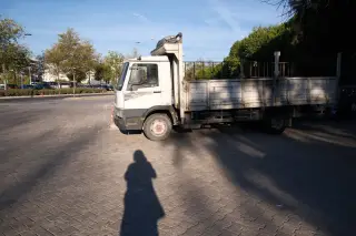 Ilustrasi: Sebuah truk bak terbuka terparkir di area berpaving dengan bayangan orang di latar depan.