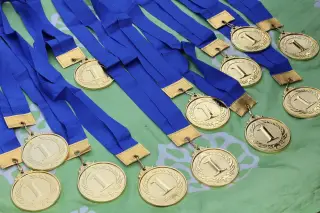 Ilustrasi: Deretan medali emas juara satu dengan pita biru tertata di atas kain hijau.