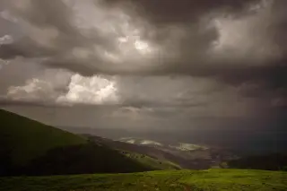Ilustrasi: Pemandangan bukit hijau di bawah langit berawan mendung yang pekat dan gelap.